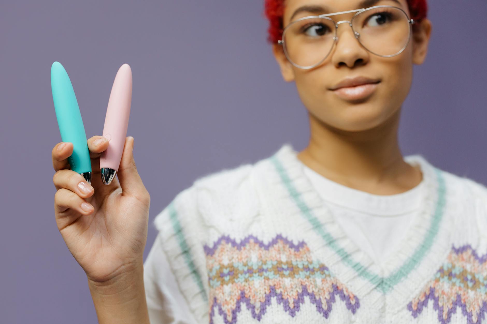 Mujer joven con gafas sosteniendo vibradores coloridos contra un fondo morado